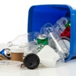 An overflowing blue recycling bin with a mix of clean recyclables and contaminating trash, symbolizing the failing US recycling system.